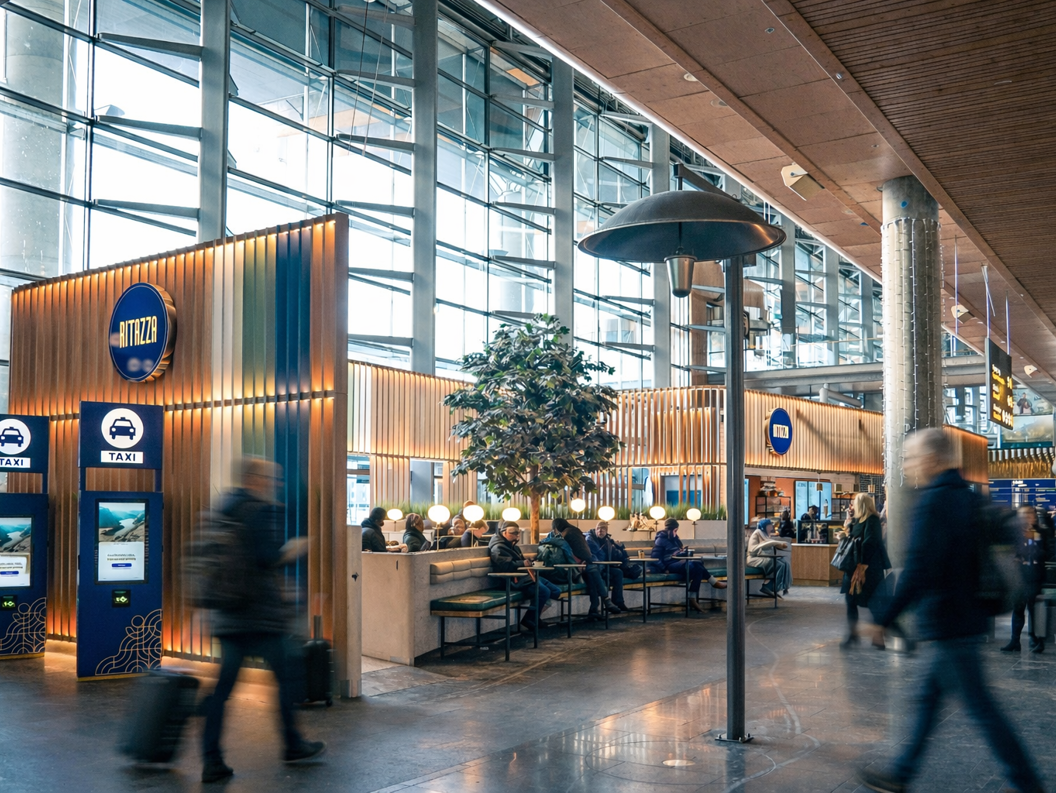 Modern coffee shop interior with decorative wooden panels, hanging lights, and a large indoor tree providing a cozy atmosphere. Patrons seated at tables with coffee and snacks on display