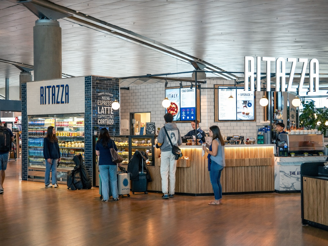 Blå Ritazza kaféskilt omgitt av grønt, med en refleksjon av et moderne interiør i glassvinduet, som antyder en koselig og innbydende atmosfære