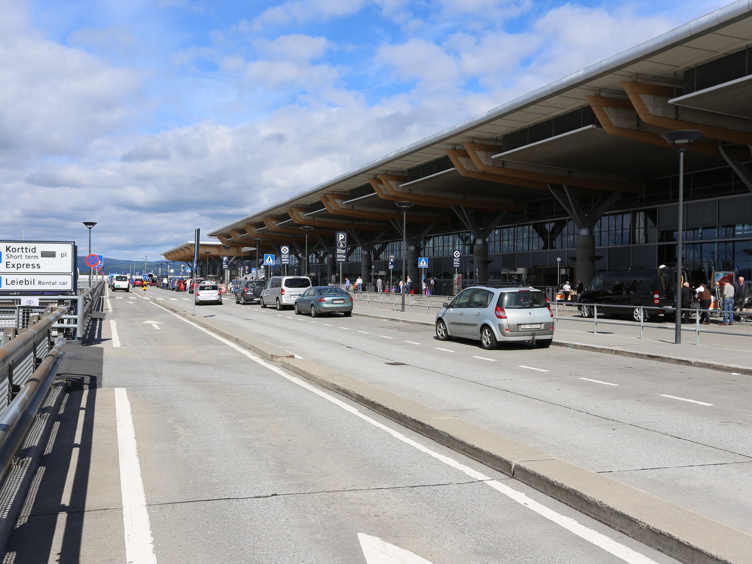 Uteområde ved en moderne flyplassterminal med biler, skilting for korttidsparkering, leiebil og reisende ved terminalinngangen