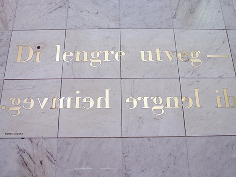 Gulletekst på et skilt montert opp-ned på en vegg med teksten "Di lengre utveg – di lengre heimveg" på norsk, designet av Kurt Johannessen. Skiltet er festet på en lys steinoverflate