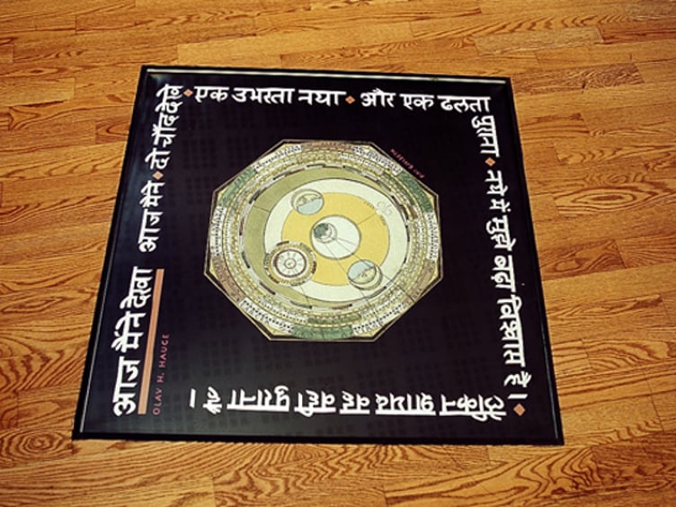 Teppe med sirkulært designelement i midten, omgitt av hvit skrift på et fremmed språk, ligger på et tregulv