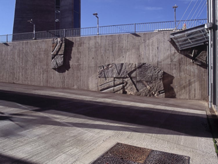 Betongvegg med abstrakt kunst og skulpturer i et urbant miljø, solrik himmel i bakgrunnen
