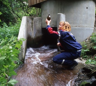 Avinor employee taking a water sample in a stream, wearing a blue rain jacket, surrounded by green vegetation.