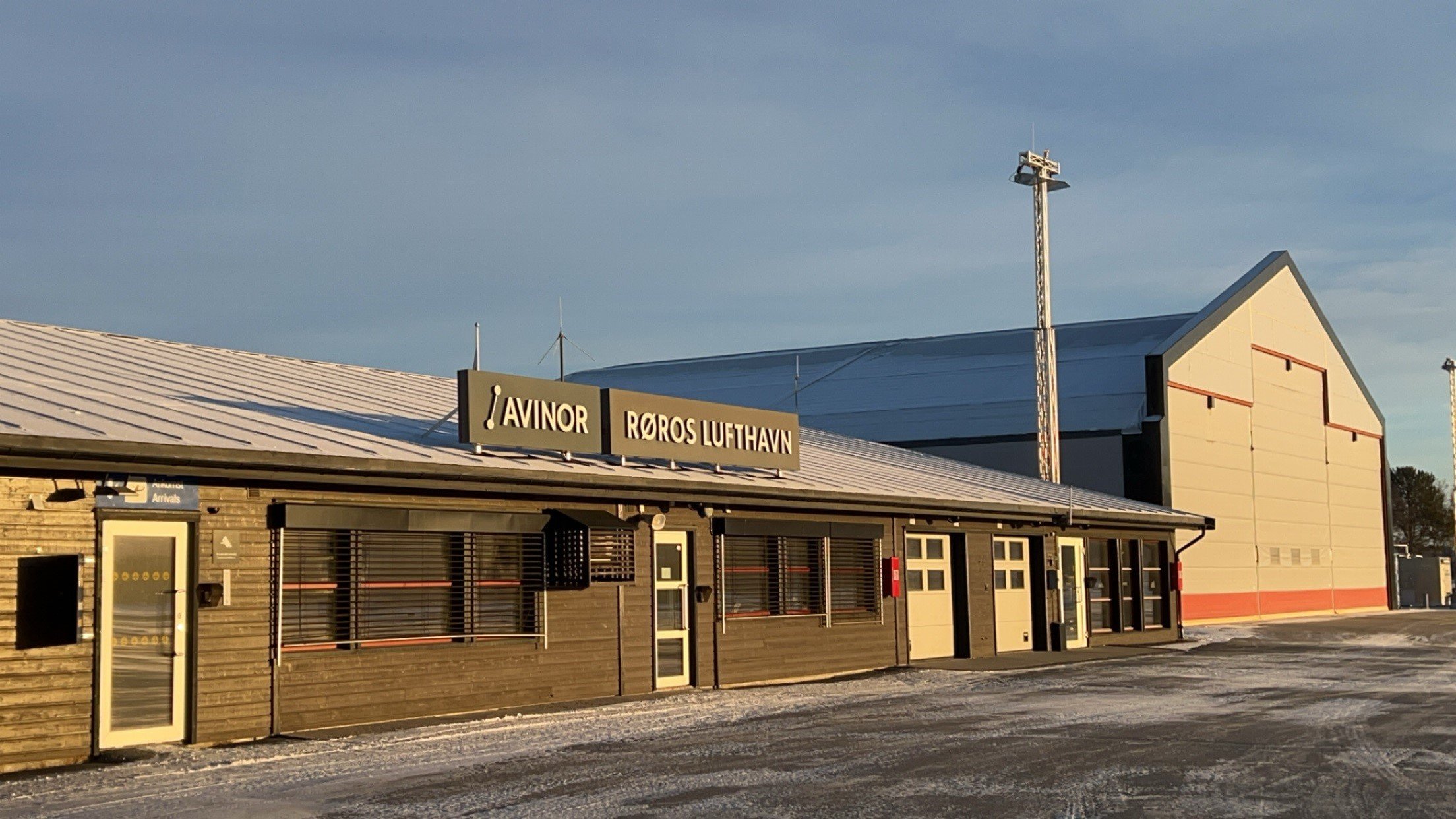 Kontrolltårn ved Røros flyplass med snø på taket, en parkeringsplass foran og en bil til venstre. Vinterlig landskap under klar himmel.
