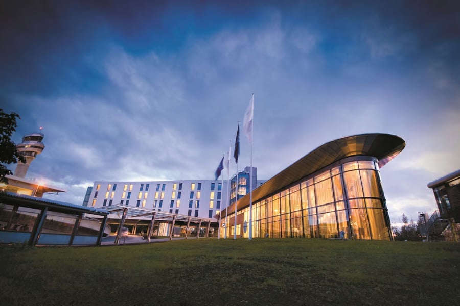 Moderne hotellbygning med glasfasade ved sidan av flyplasstårn i skumringa, omgjeven av grønt gras.