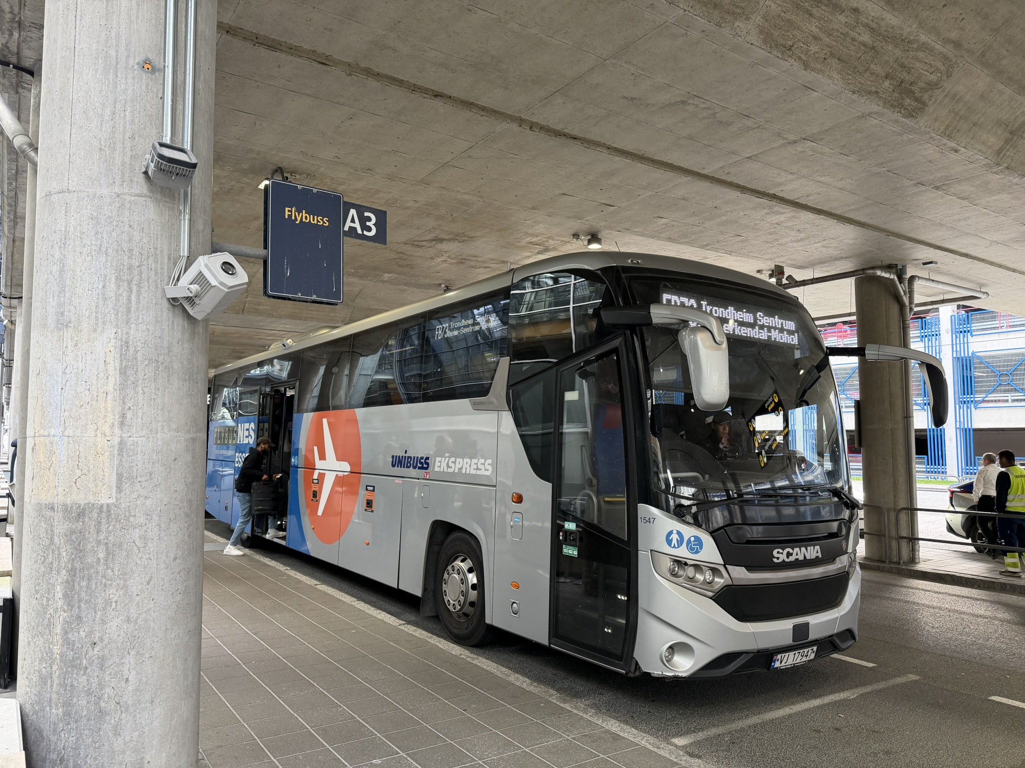 Blue bus outside an airport terminal in Norway, with passengers disembarking and some standing with luggage. Modern building and parking lot in the background. Gray sky and humid weather.