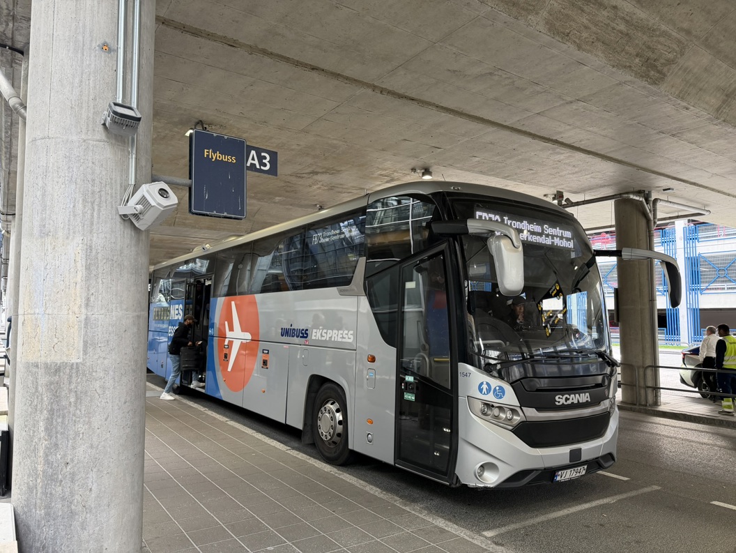 Blue bus outside an airport terminal in Norway, with passengers disembarking and some standing with luggage. Modern building and parking lot in the background. Gray sky and humid weather.