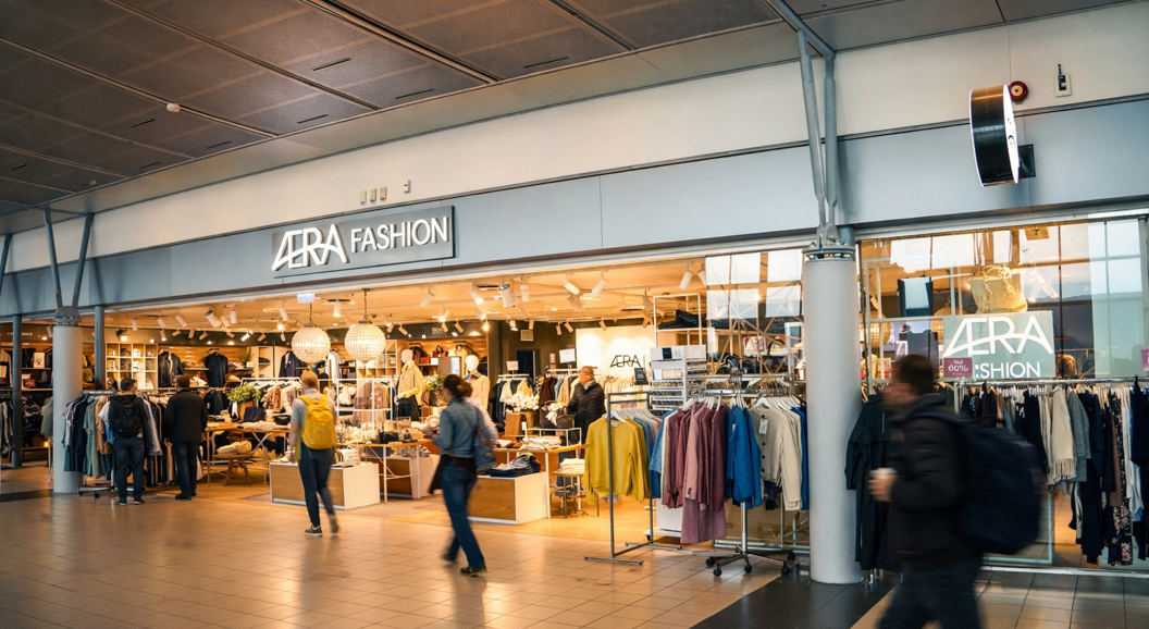 Motebutikkens inngang viser stilige mannequiner og tilbehør, med fremtredende "AERA Fashion" skilt over. Interiøret viser et utvalg av klær og smykker under varm belysning