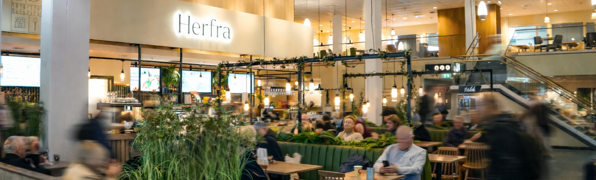Busy modern café inside an airport with passengers seated at tables, warm lighting, and the Herfra sign visible above the counter