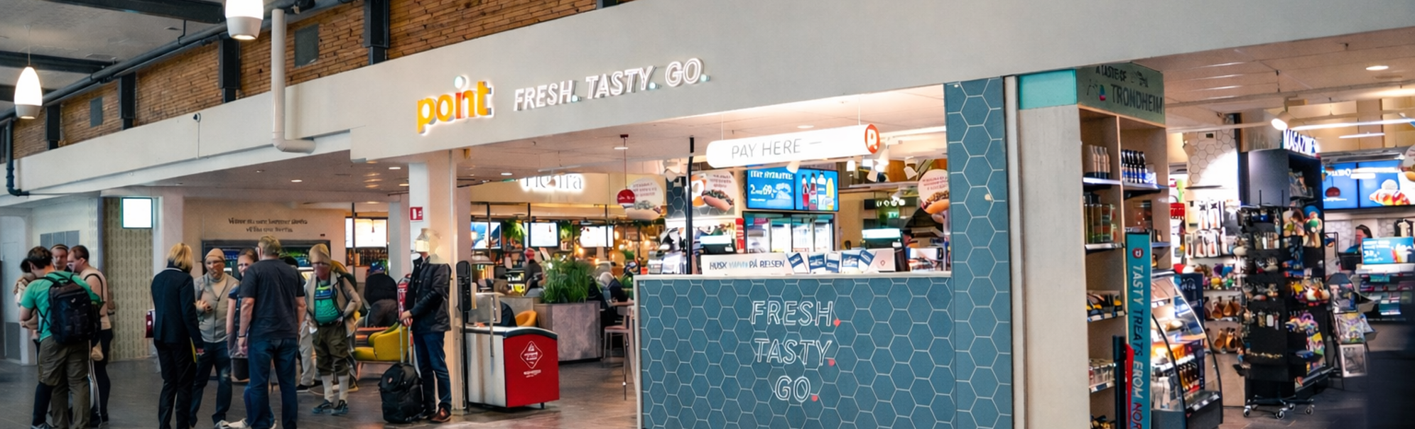 Flyplassmatkrok og nærbutikk med reisande som ventar i kø nær ein Point Fresh Tasty Go-disk, innandørs terminalsceanne