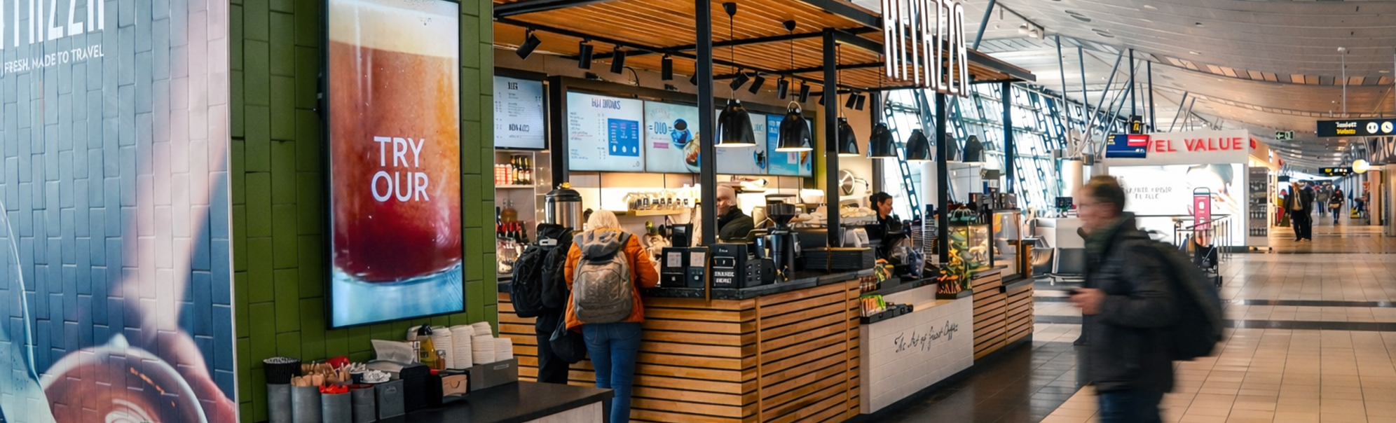 Flyplassterminal-kiosk for kaffi og bakverk med reisande som går forbi under moderne, bogeforma taklys