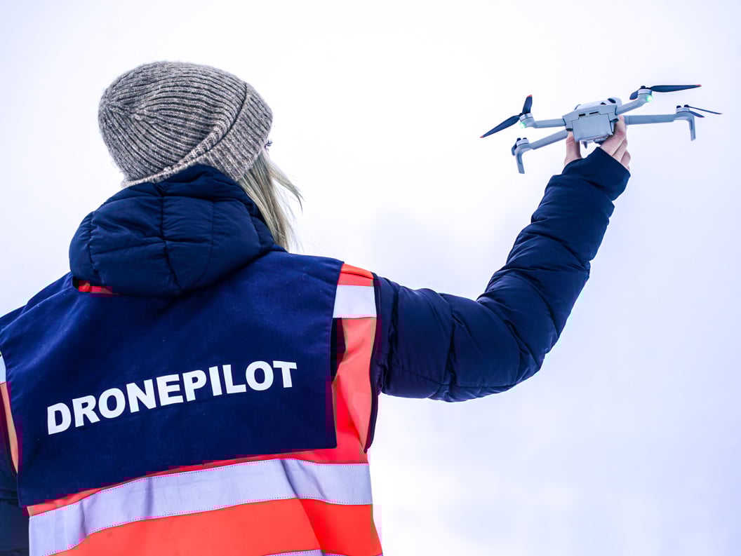 Kvinne i oransje jakke med dronepilot-vest som held ein drone i handa opp mot himmelen