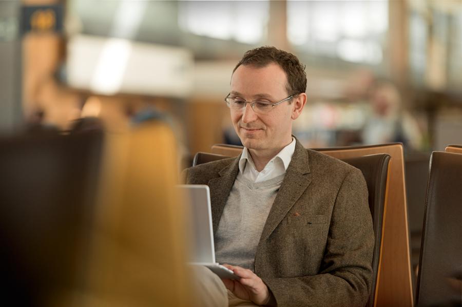 A man with short brown hair and glasses sitting in one of the seats by a gate, looking at his laptop on his lap.